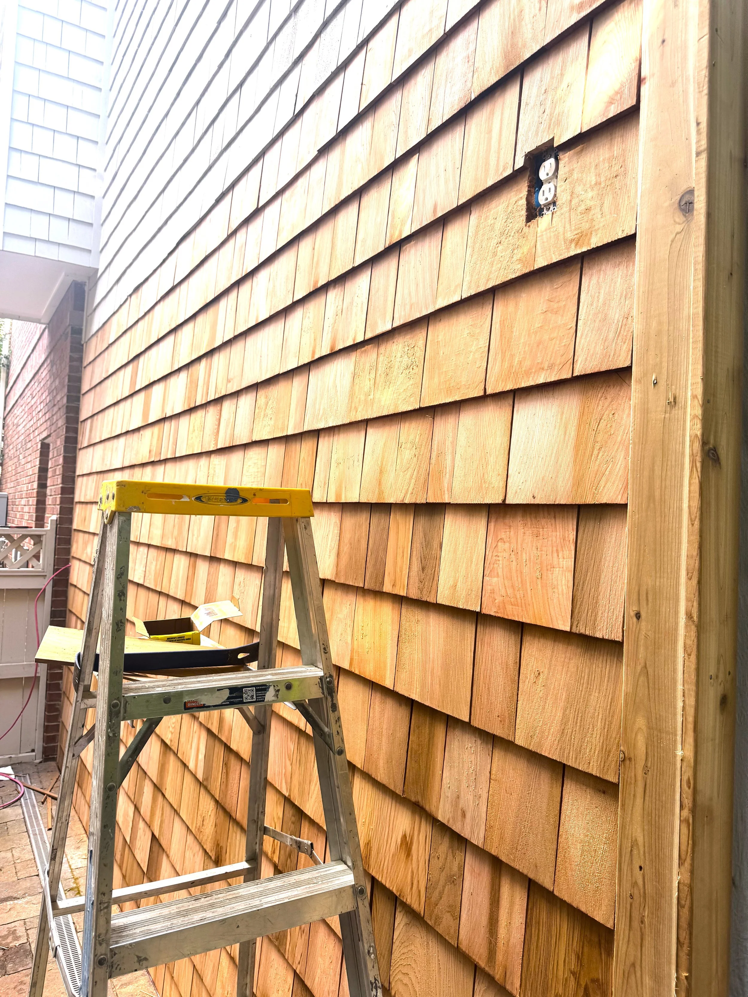 A wooden exterior wall under construction with new wooden shingles, a step ladder, and an electrical outlet box installed in the wall.