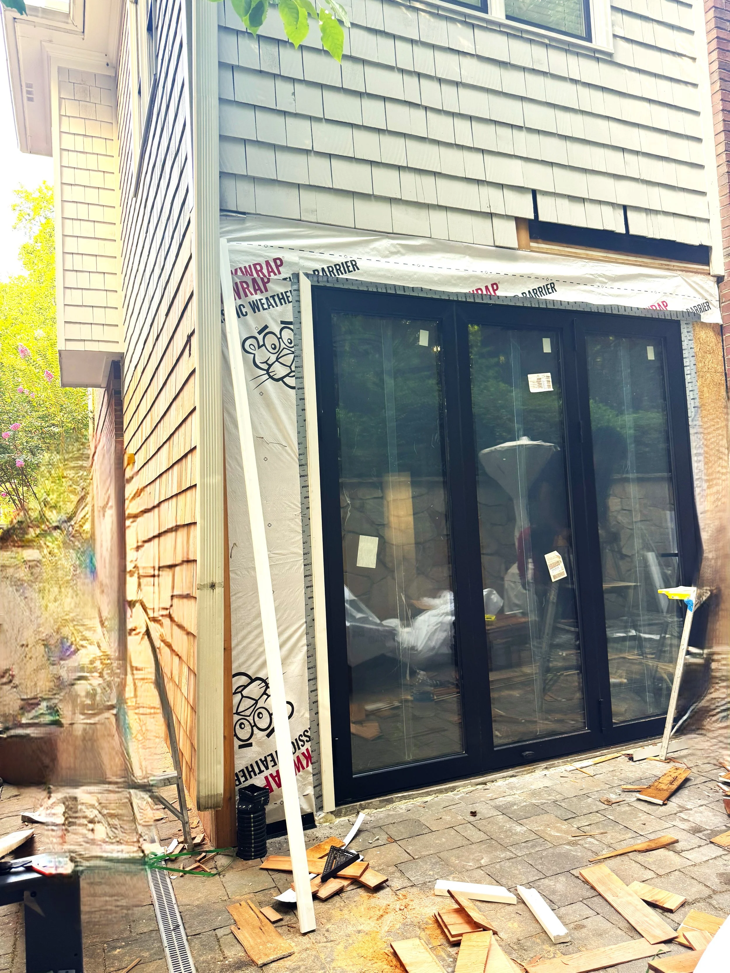 Part of a house under construction with a sliding glass door, weatherproofing wrap, and damaged wooden siding debris on the brick-paved ground.