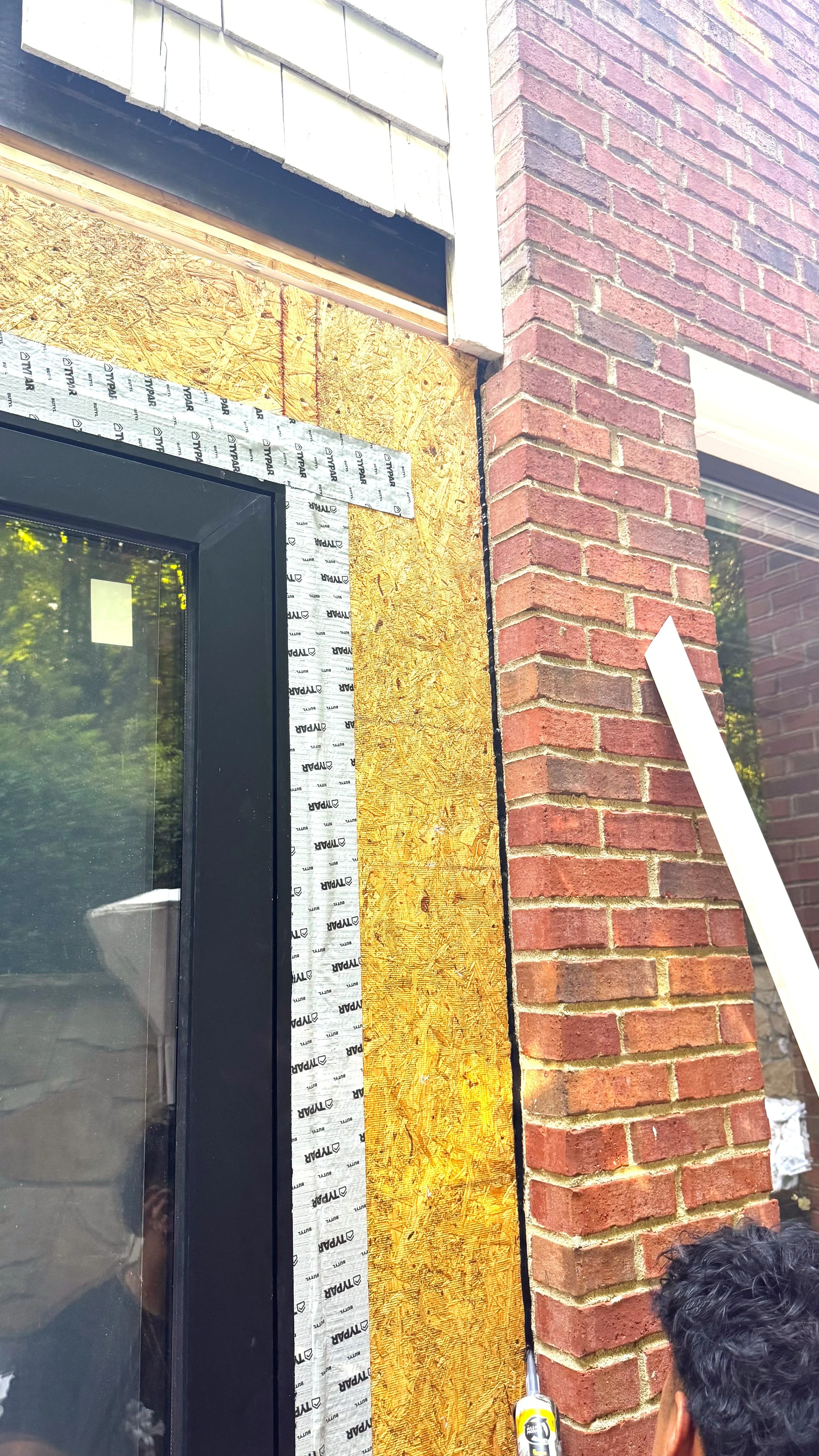 Person using a caulking gun on a brick wall next to a window with construction materials around.