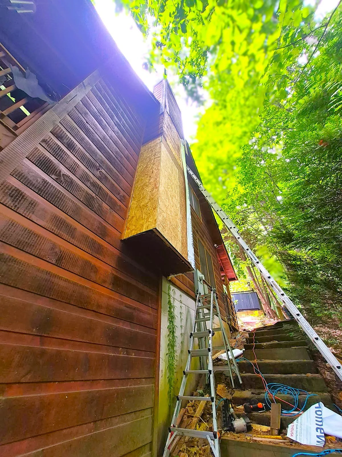 Exterior of a wooden house under construction, with a ladder leaning against it and construction tools on the ground. The house has horizontal wooden siding and is surrounded by trees with green leaves.