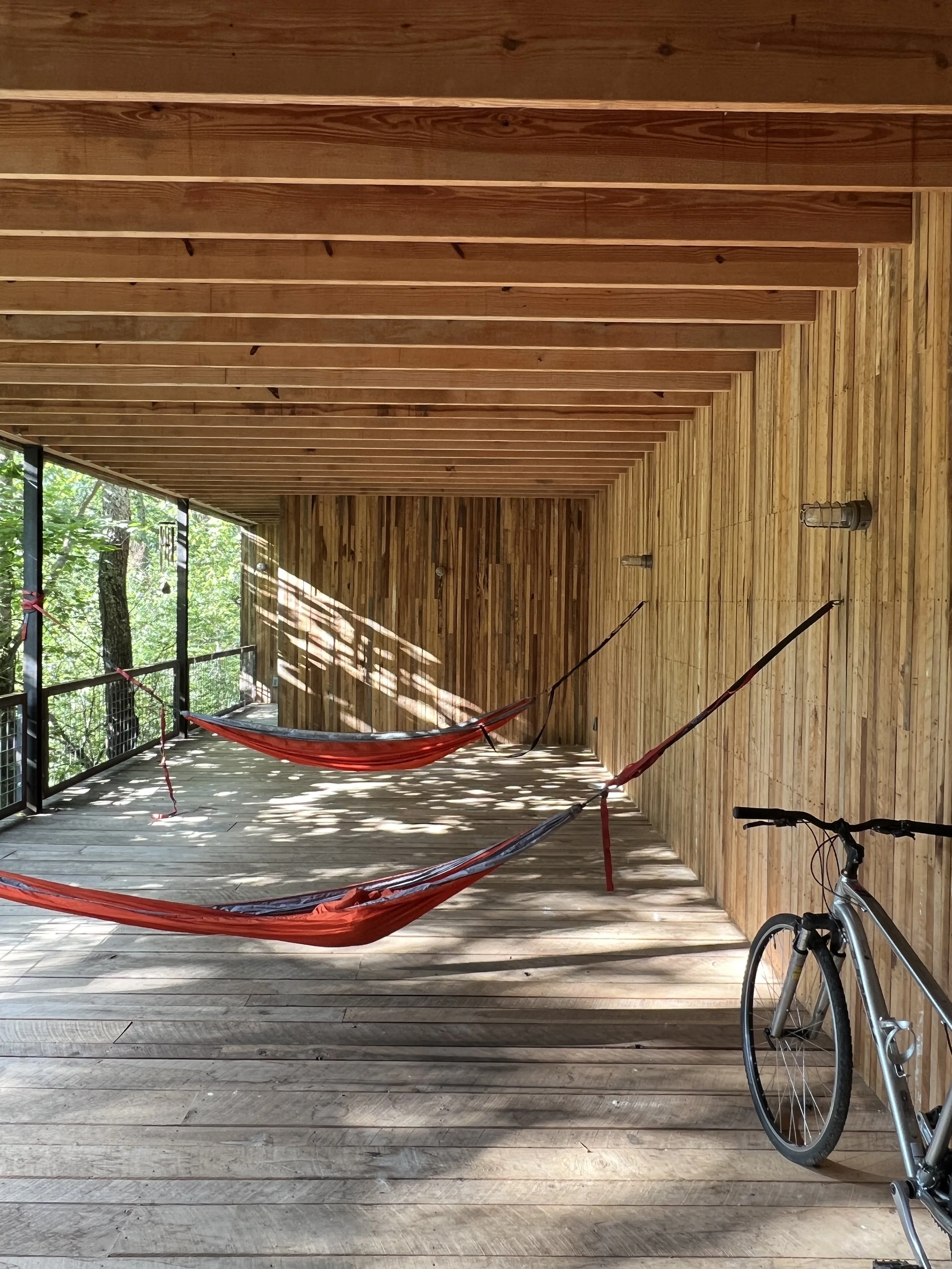 A wooden balcony with a red hammock and a bicycle in the corner, surrounded by trees visible through the railing.