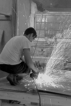 A person crouching and welding metal, with sparks flying around in a workshop or construction area.