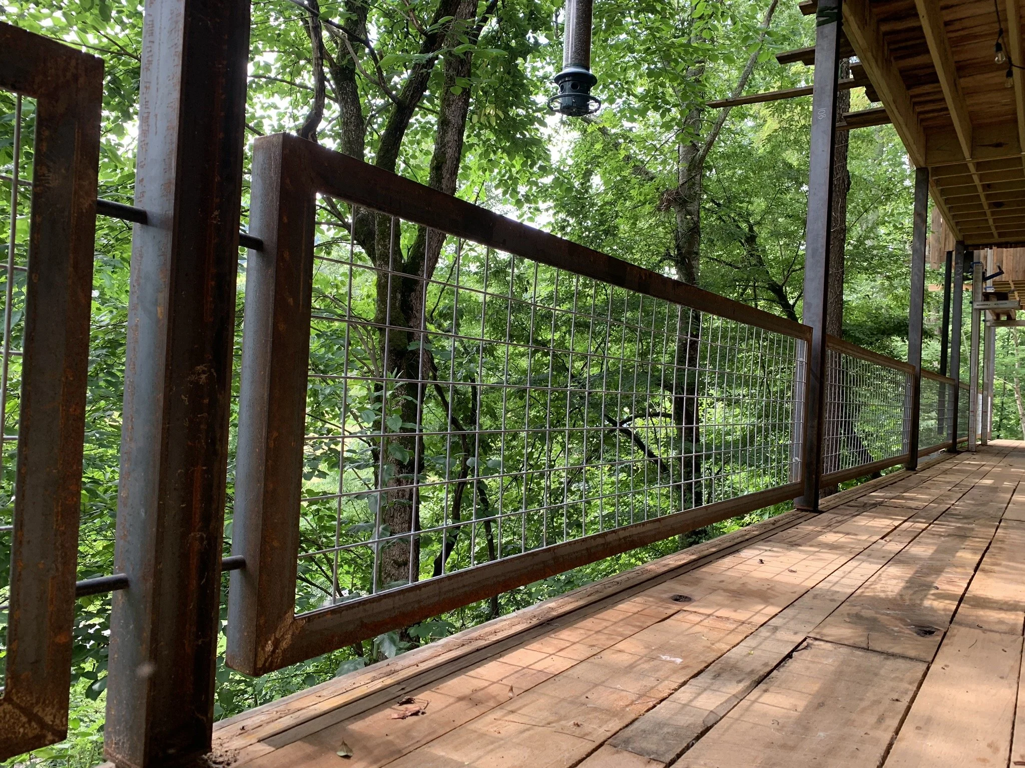 Wooden porch overlooking trees in a forest, with a wire fence and a metal post with a bird feeder hanging from it.
