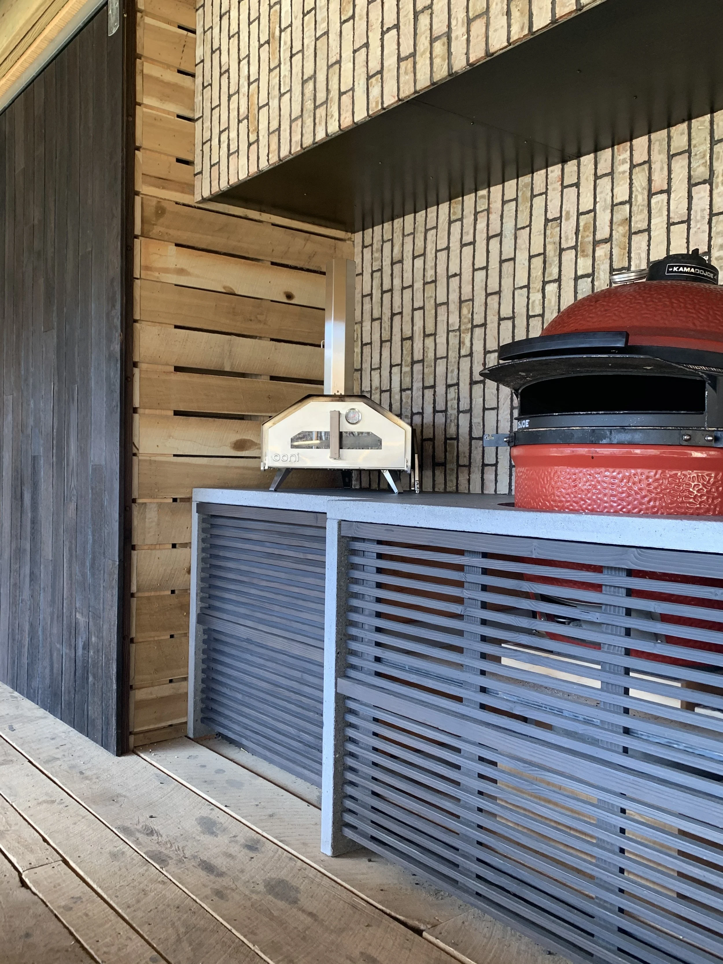 A backyard patio area with a red Kamado-style barbecue grill, a gray wooden fence, and a brick wall with a black overhang. There is also a small white metal oven or smoker on a raised surface.