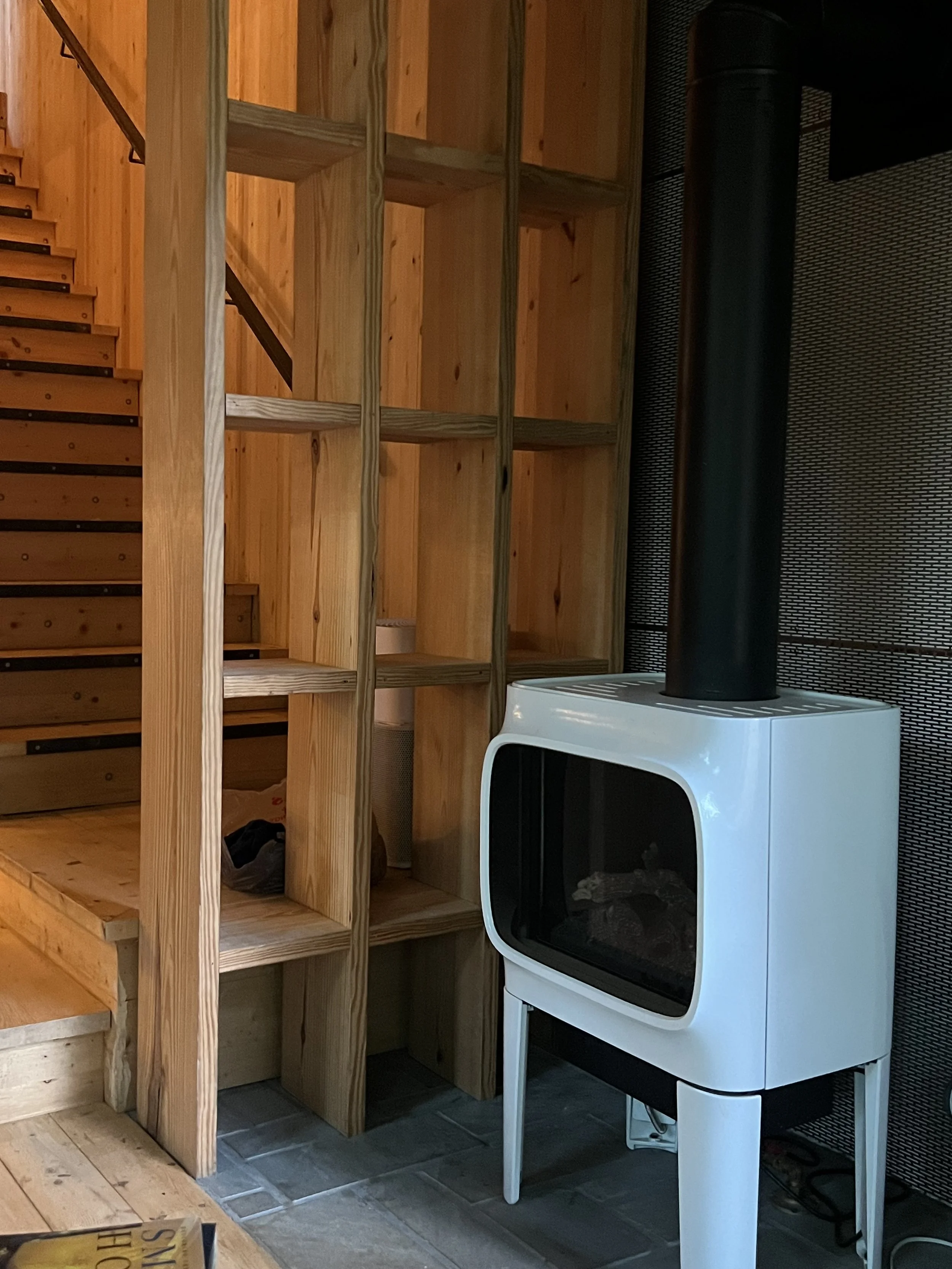 A small, white, modern fireplace with a glass front, placed on a white stand with four legs, located next to a wooden shelf with multiple compartments in a room with black tile flooring.