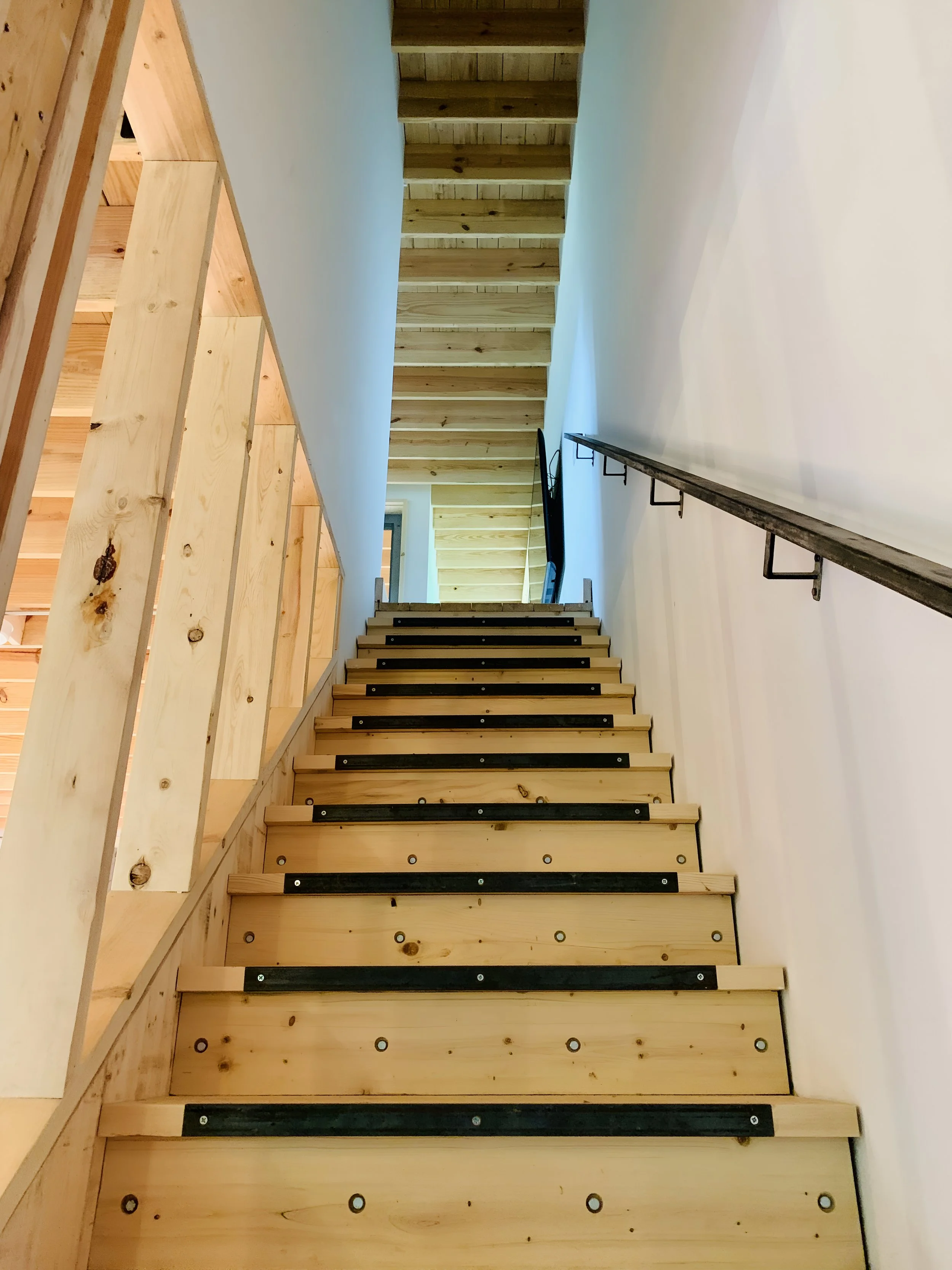 Unfinished wooden staircase with black anti-slip strips on each step, metal handrail on the right, and a partial view of wooden wall paneling on the left.