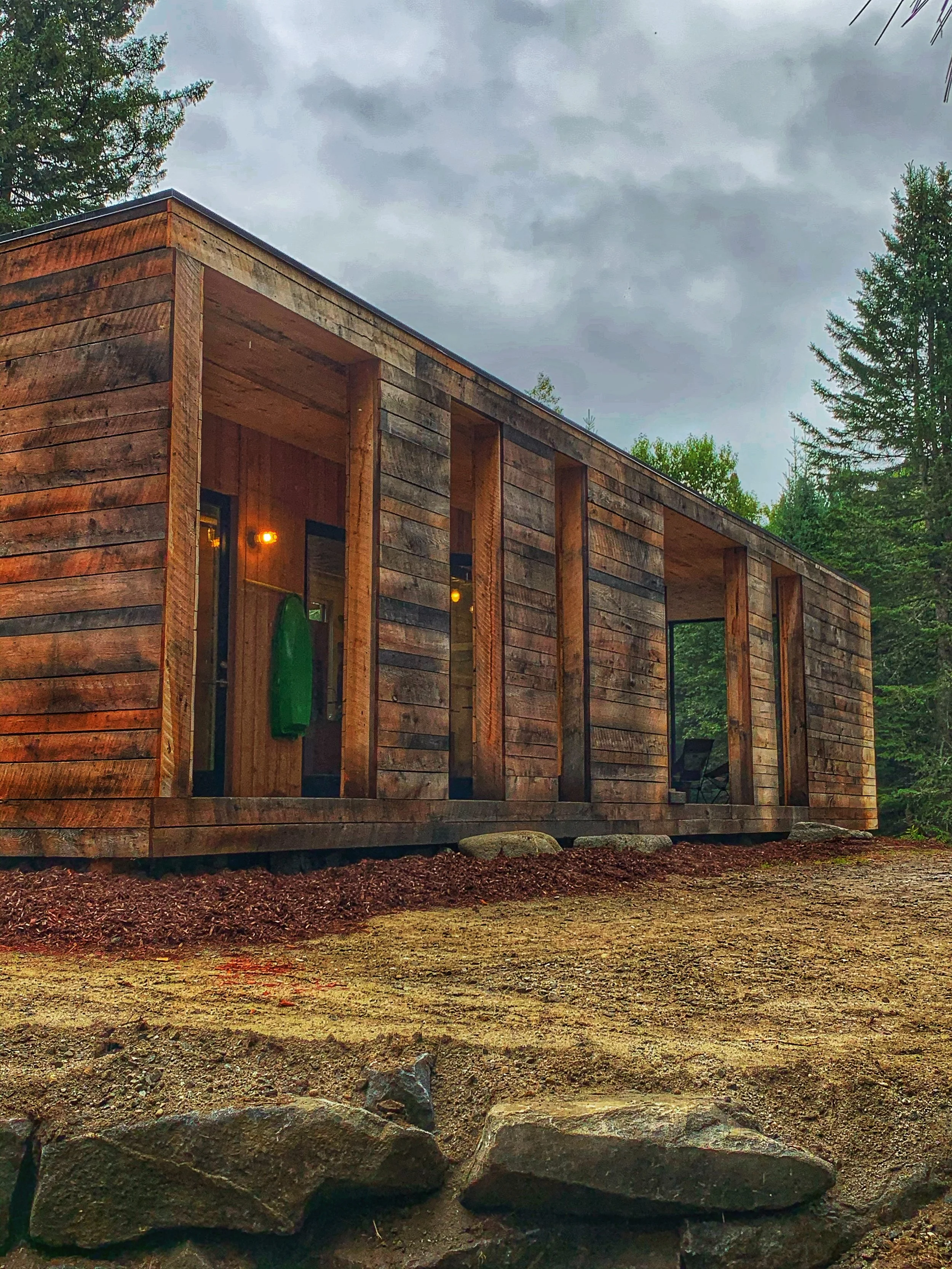 A modern house under construction with wooden exterior walls and large windows, surrounded by trees, on a cloudy day.