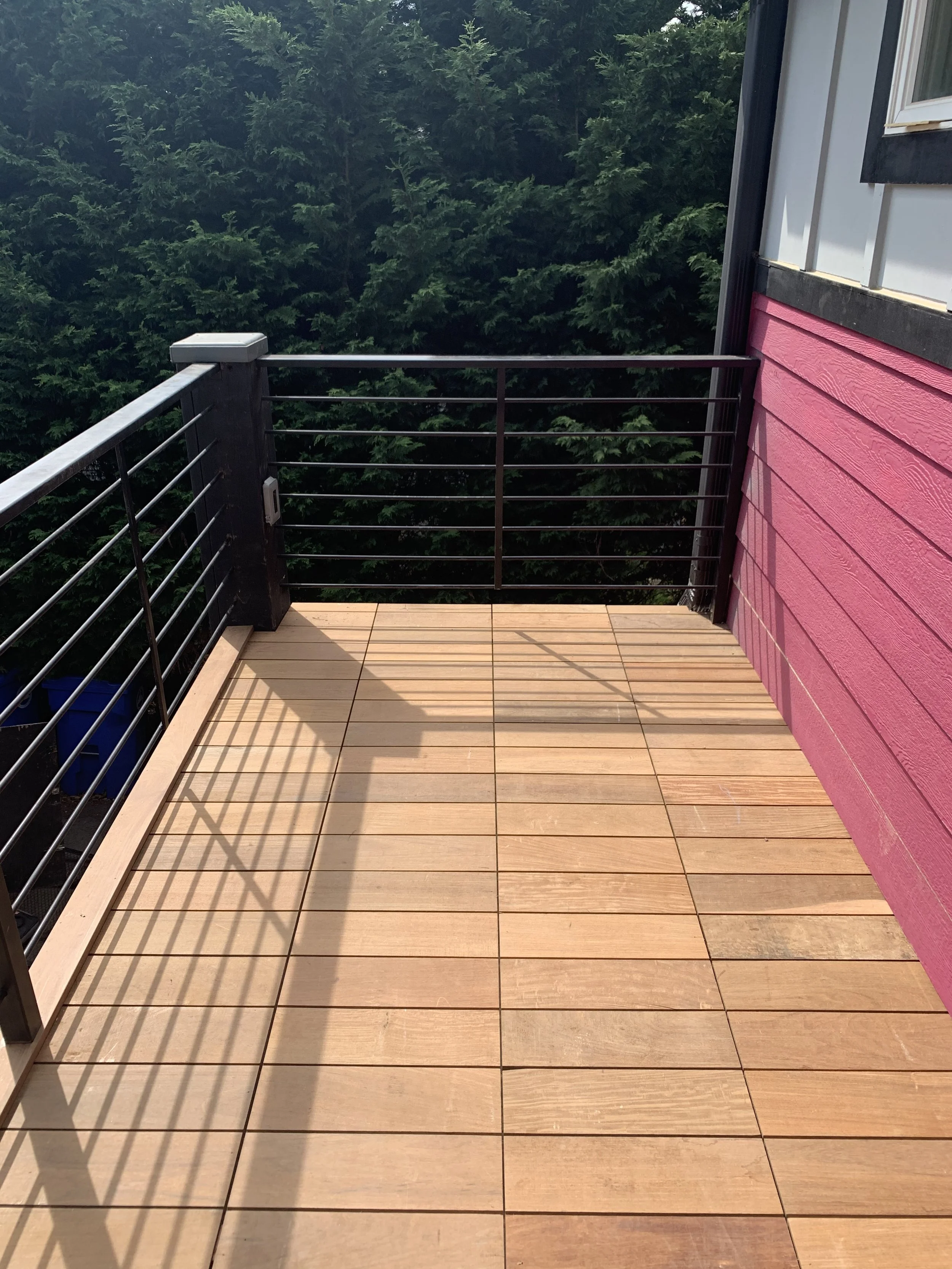 View from a balcony with wooden flooring, black metal railing, pink siding on the house, and green trees in the background.