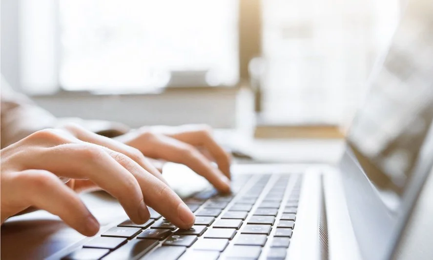Close-up of a person typing on a laptop keyboard with a bright window in the background