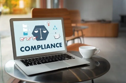Laptop displaying the word 'Compliance' with icons of a balance scale, lightbulb, checklist, and paper, on a round table next to a coffee cup in a modern office setting.