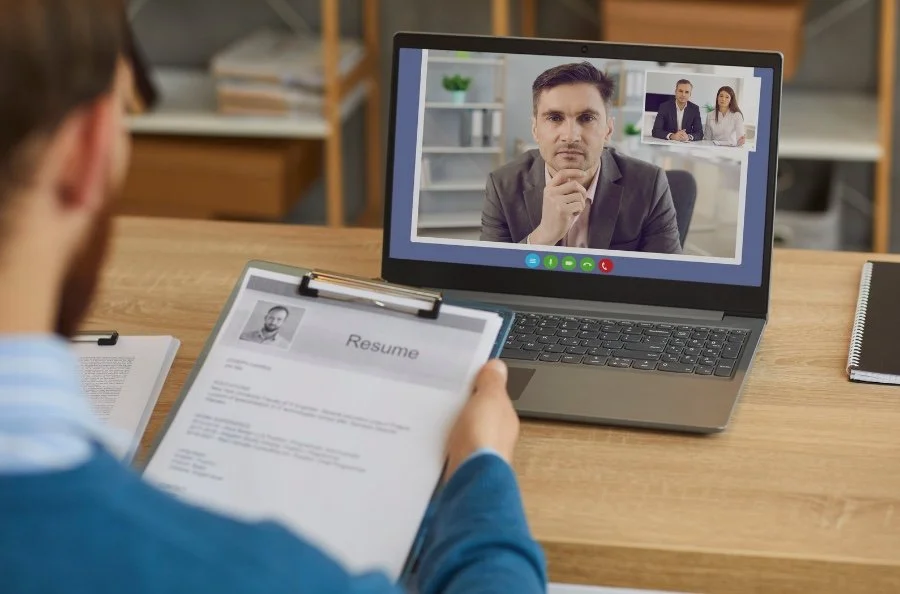 Person participating in a virtual job interview with a recruiter on a laptop computer, holding a resume in a well-lit office.