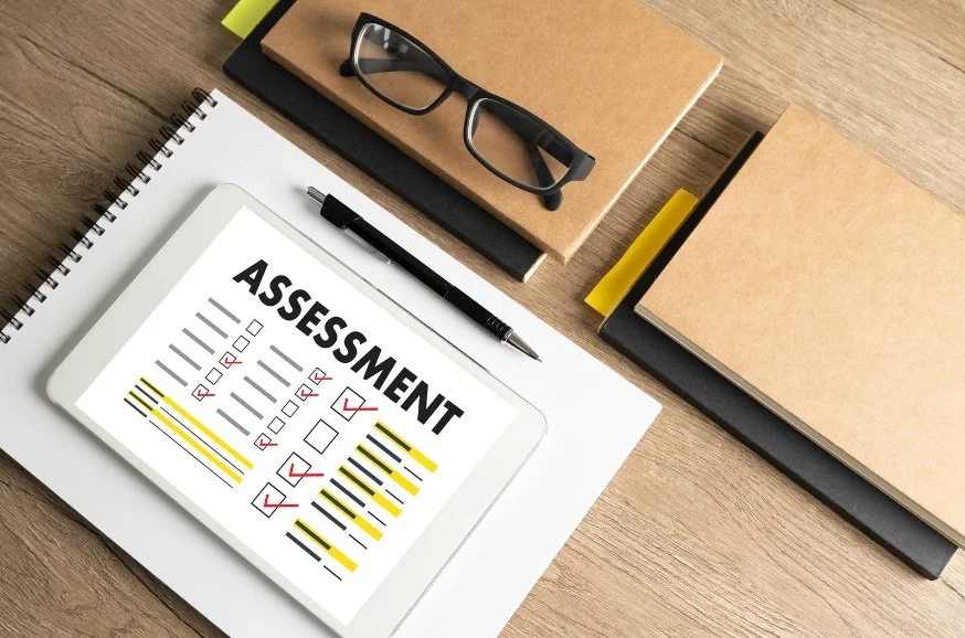 Digital tablet displaying an assessment form with checkboxes and yellow highlights on a wooden desk surrounded by notebooks, a pen, and a pair of glasses.
