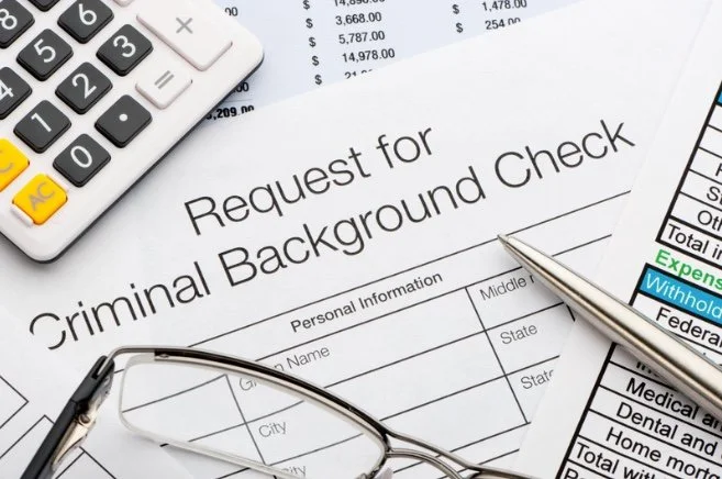 A close-up of a criminal background check request form, a pen, glasses, a calculator, and some documents on a desk.
