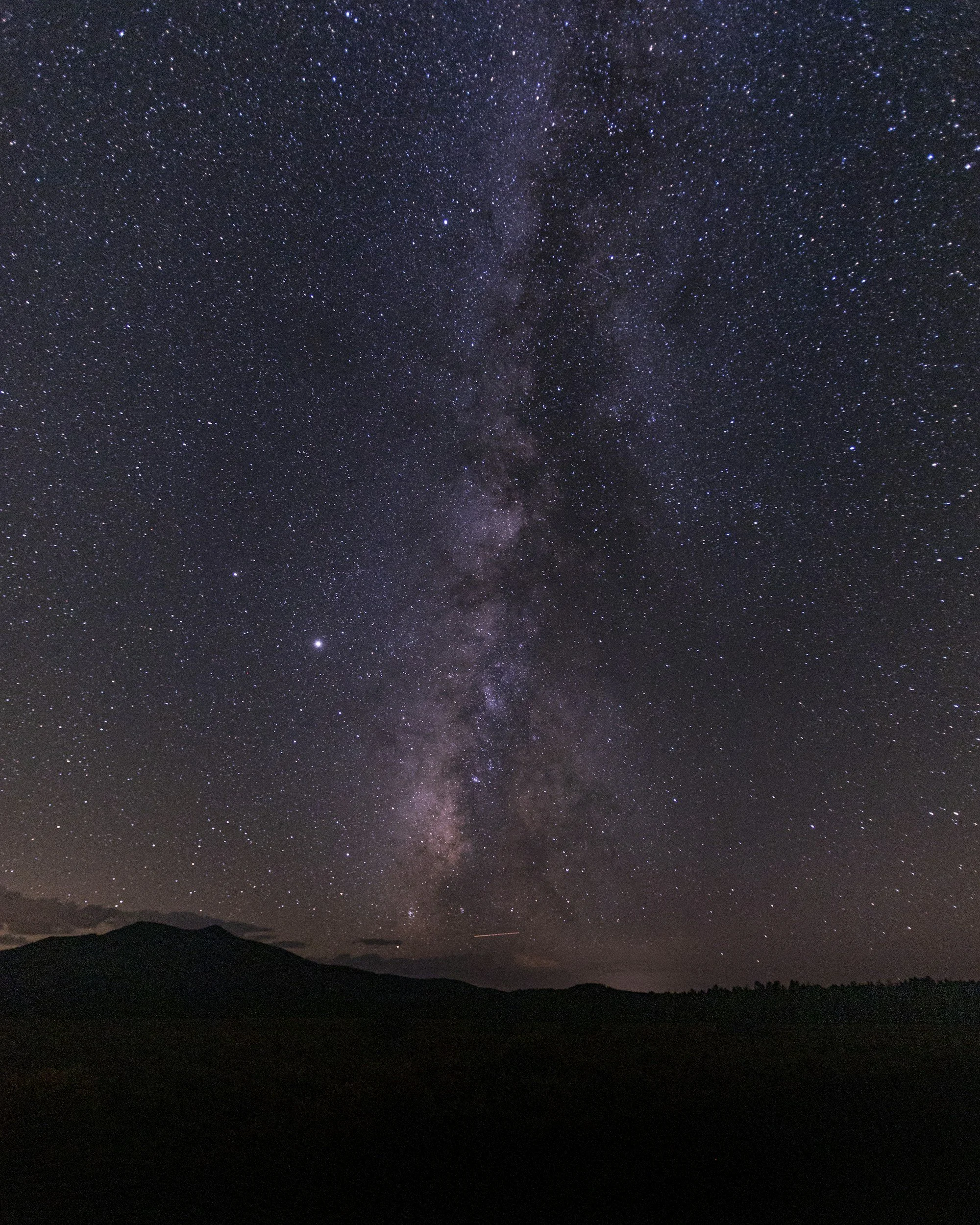Night sky filled with stars and the Milky Way galaxy visible above a silhouette of mountains and a distant horizon.