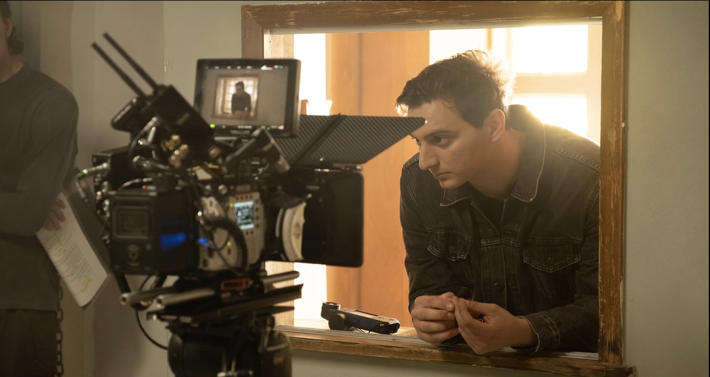 A young man with curly hair in a black denim jacket leans forward, seen through a mirror, as a camera captures his image. The background is brightly lit with sunlight, and a woman holding papers is visible on the left side.