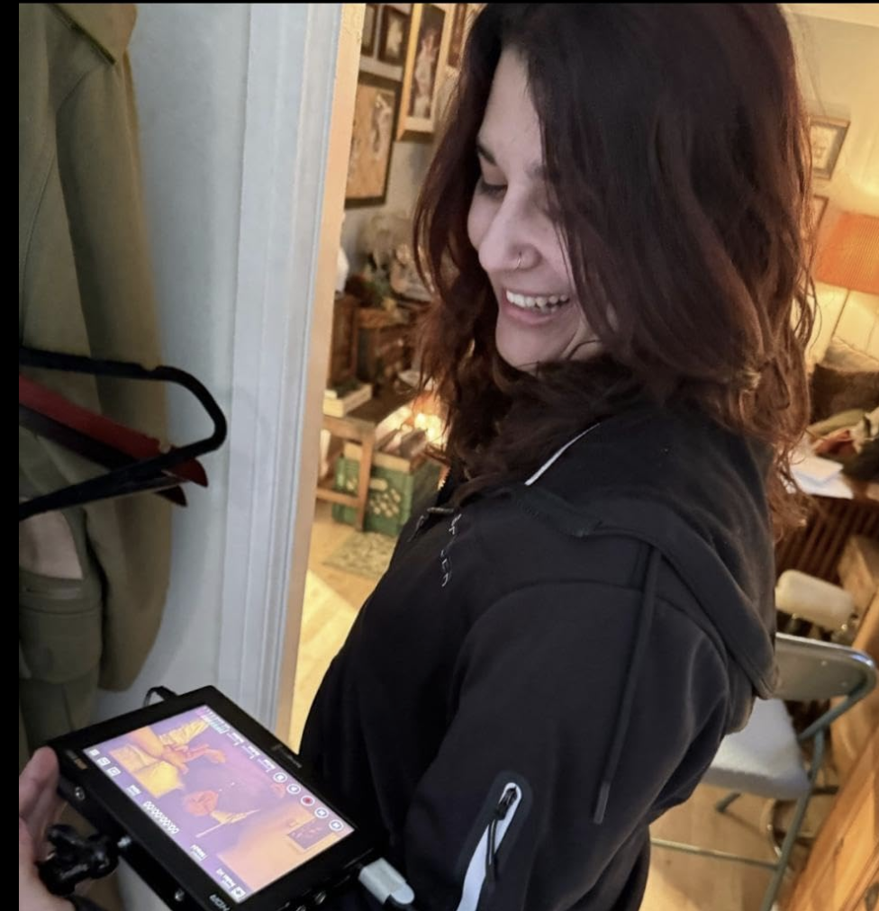A woman with shoulder-length curly brown hair and a nose piercing smiling as she looks at a handheld electronic device, possibly a camera or monitor, inside a cozy home with framed pictures on the wall and a living room in the background.
