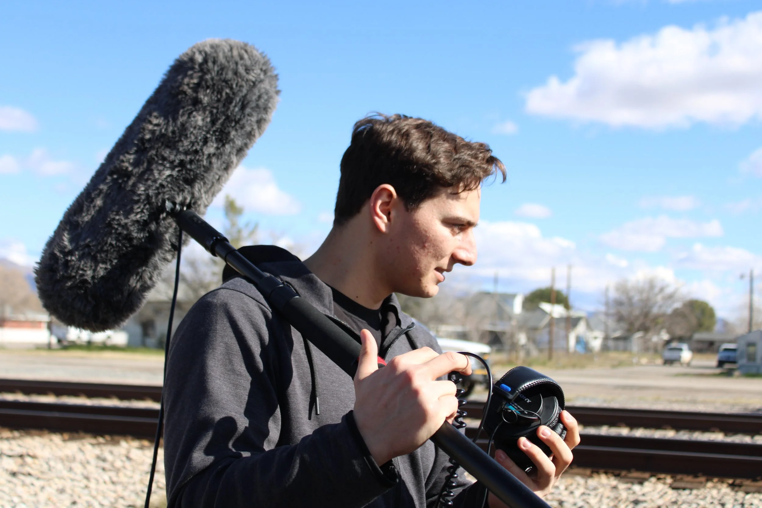 A young man with short brown hair operating a video camera with a large furry microphone attached, outdoors near train tracks under a partly cloudy sky.