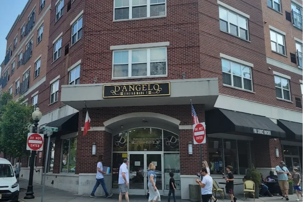 People walking past the entrance of a building with a sign that reads 'D' ANGELO Italian Market'. The building is brick with fluorescent lights and large glass doors, and there are street signs indicating 'Do Not Enter'.