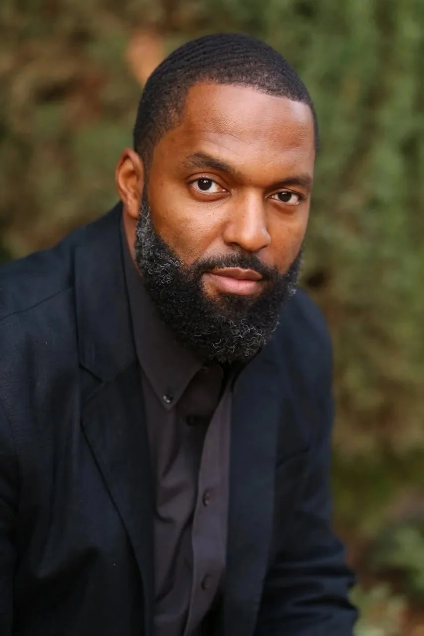 Portrait of a man with short hair and a beard, wearing a black jacket and shirt, outdoors with a blurred natural background.