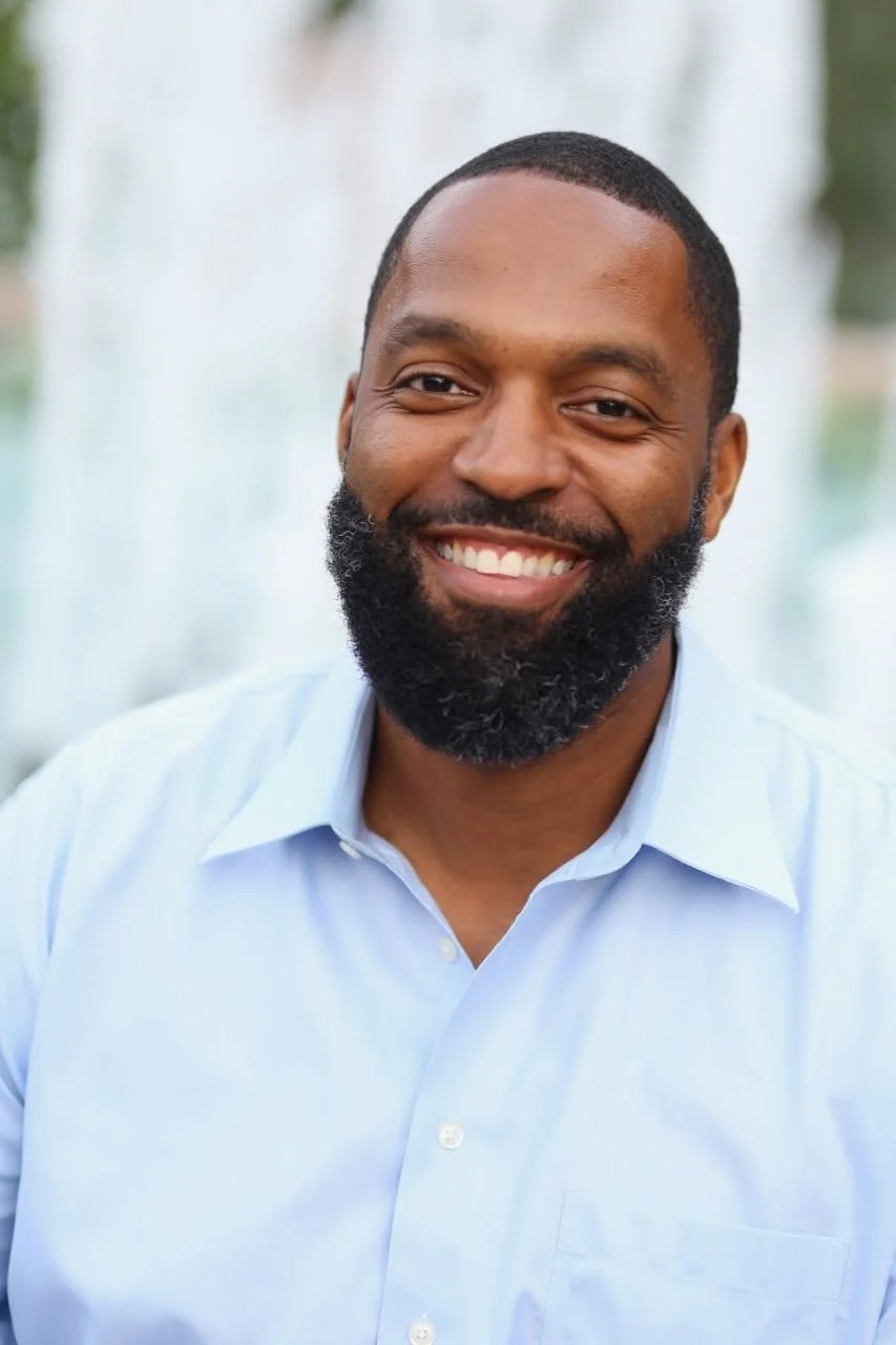 Portrait of a smiling Black man with a beard and short hair, wearing a light blue dress shirt, outdoors near a water fountain.