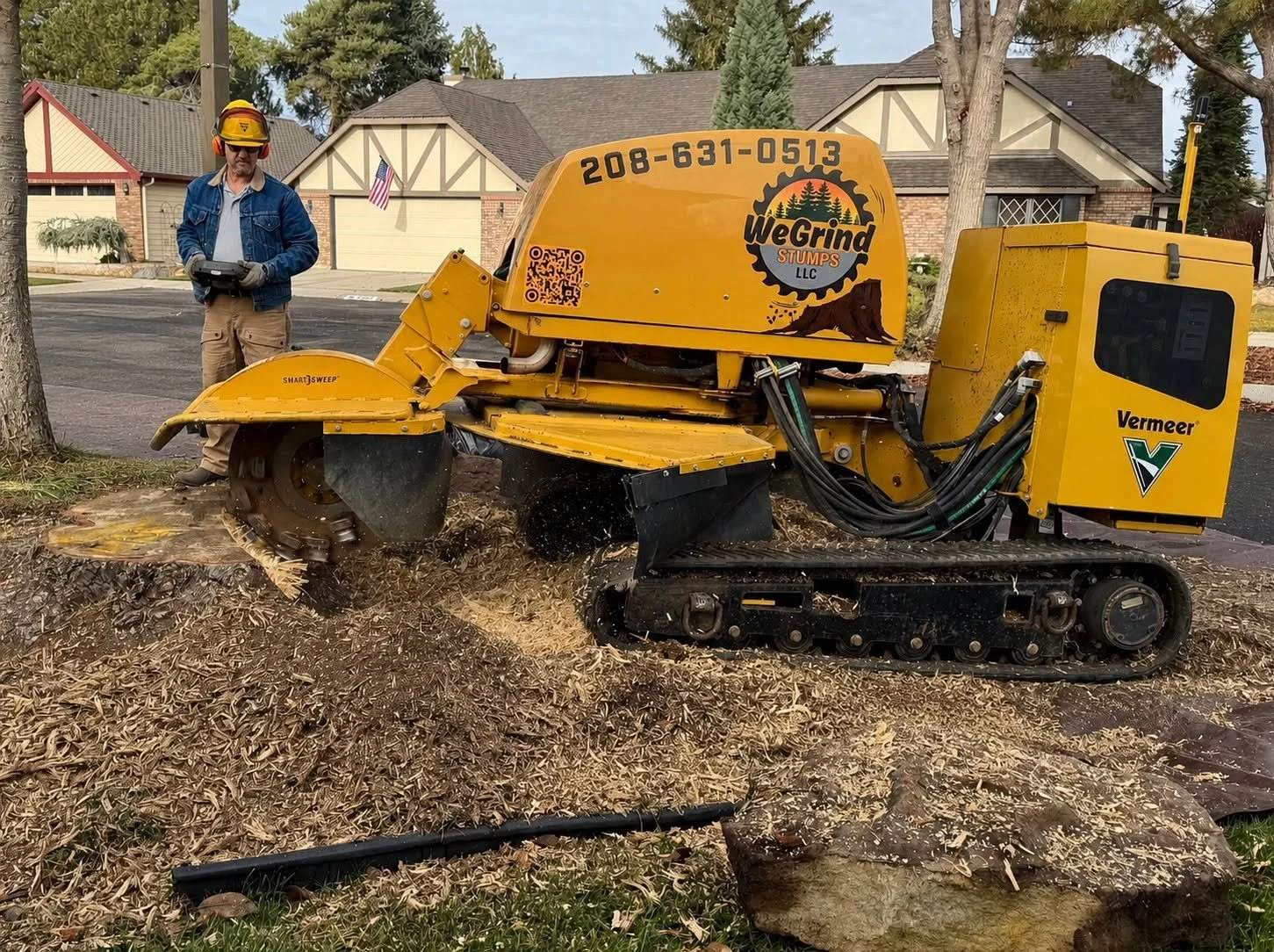 A worker operating a yellow WeGrind tree-stump grinder in a residential neighborhood, with houses and trees in the background.