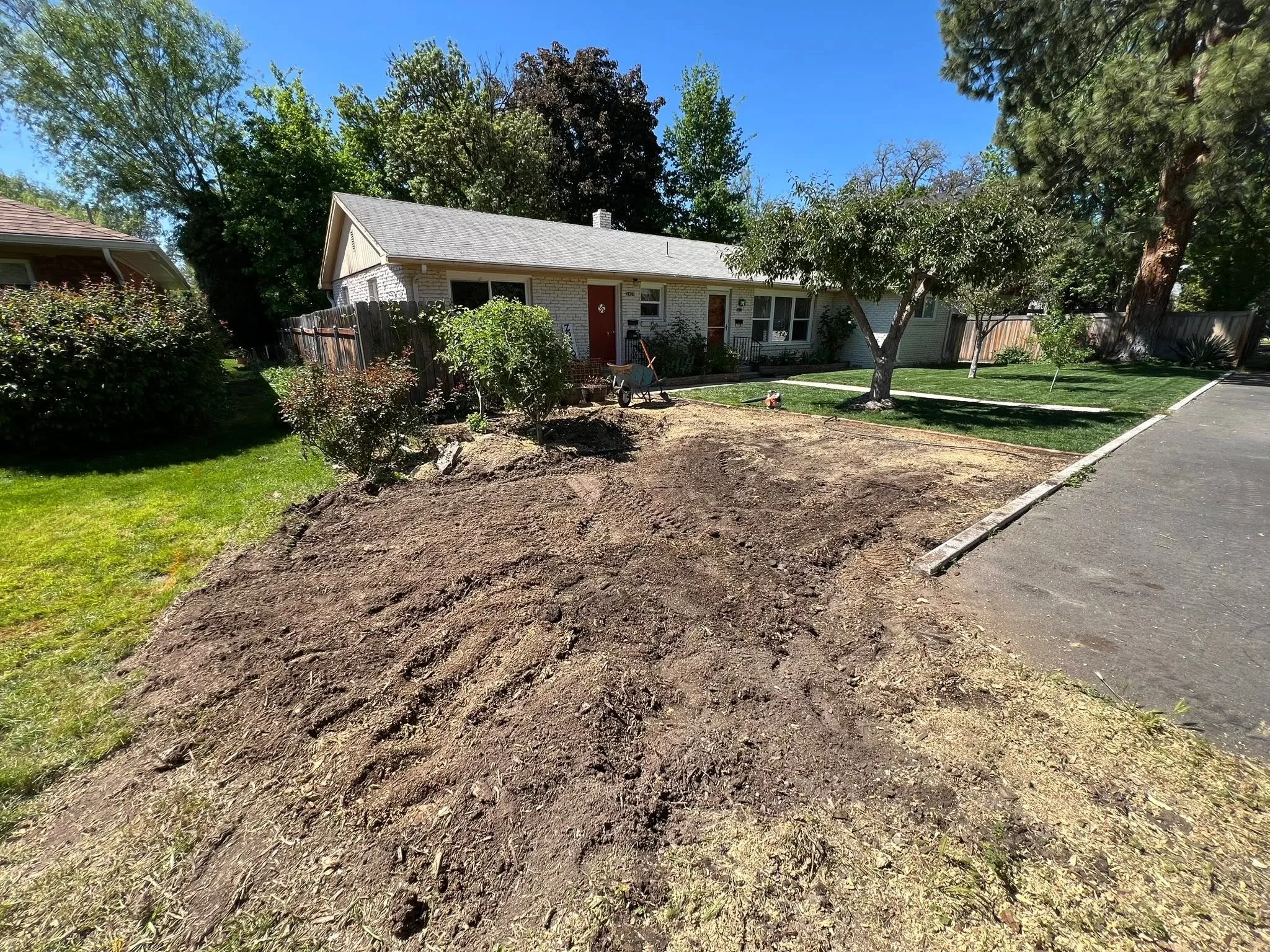 The image shows a residential backyard with a house, trees, a lawn, and a dirt patch in the foreground, indicating a clean and complete stump-grinding service.