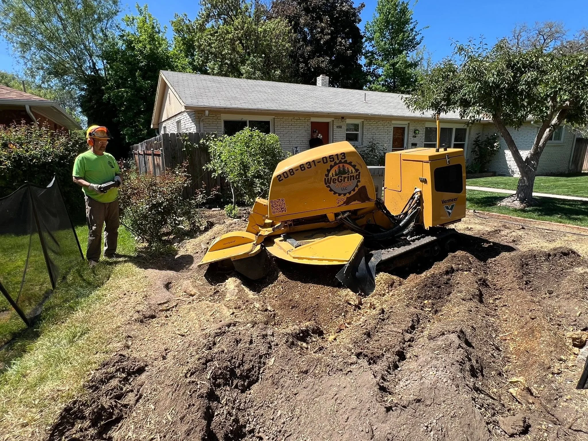 A construction worker wearing a green shirt, safety glasses, and a helmet standing next to a yellow trencher in a backyard. The house is in the background, along with trees, bushes, and a clear blue sky.