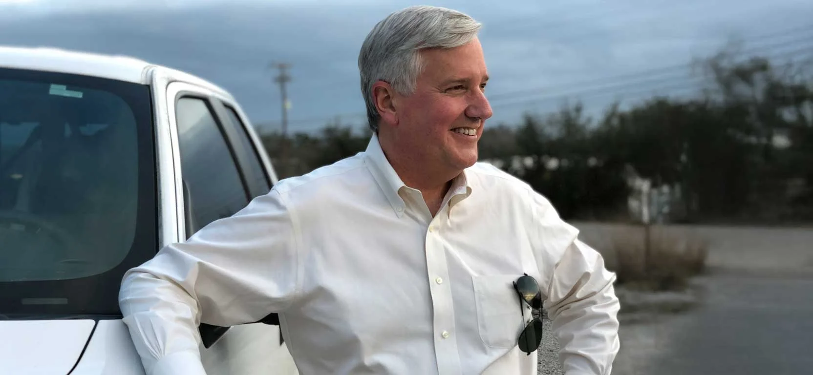 A smiling middle-aged man with gray hair wearing a white dress shirt, standing outdoors next to a parked vehicle, with trees and power lines in the background.