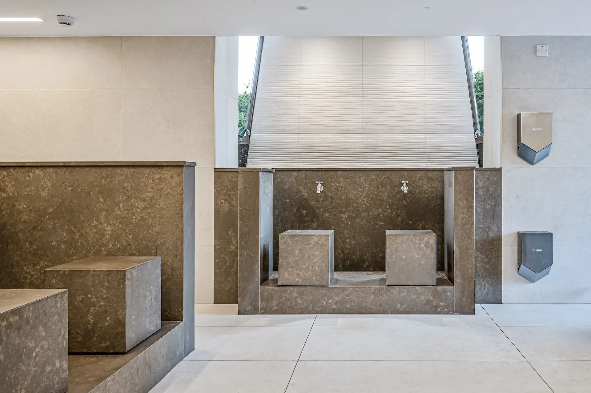 Modern public restroom with two stone sinks, faucets, benches, window shade, and Dyson hand dryers