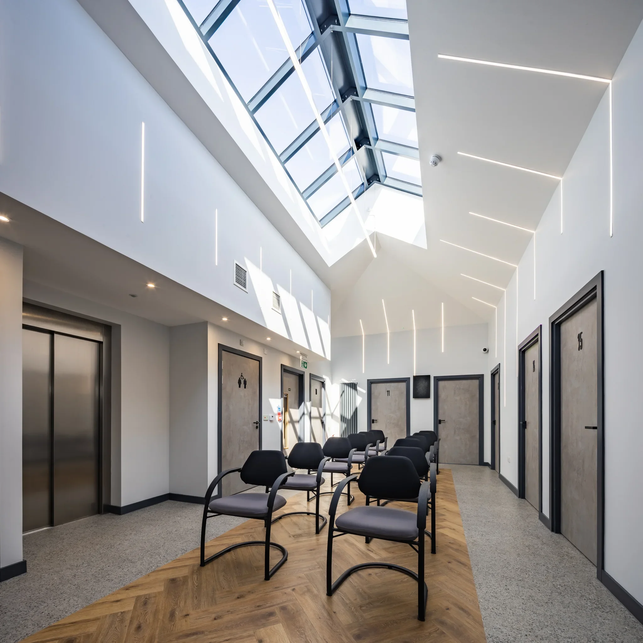 Modern clinic or office waiting area with black chairs, elevator, numbered doors, and a bright glass skylight