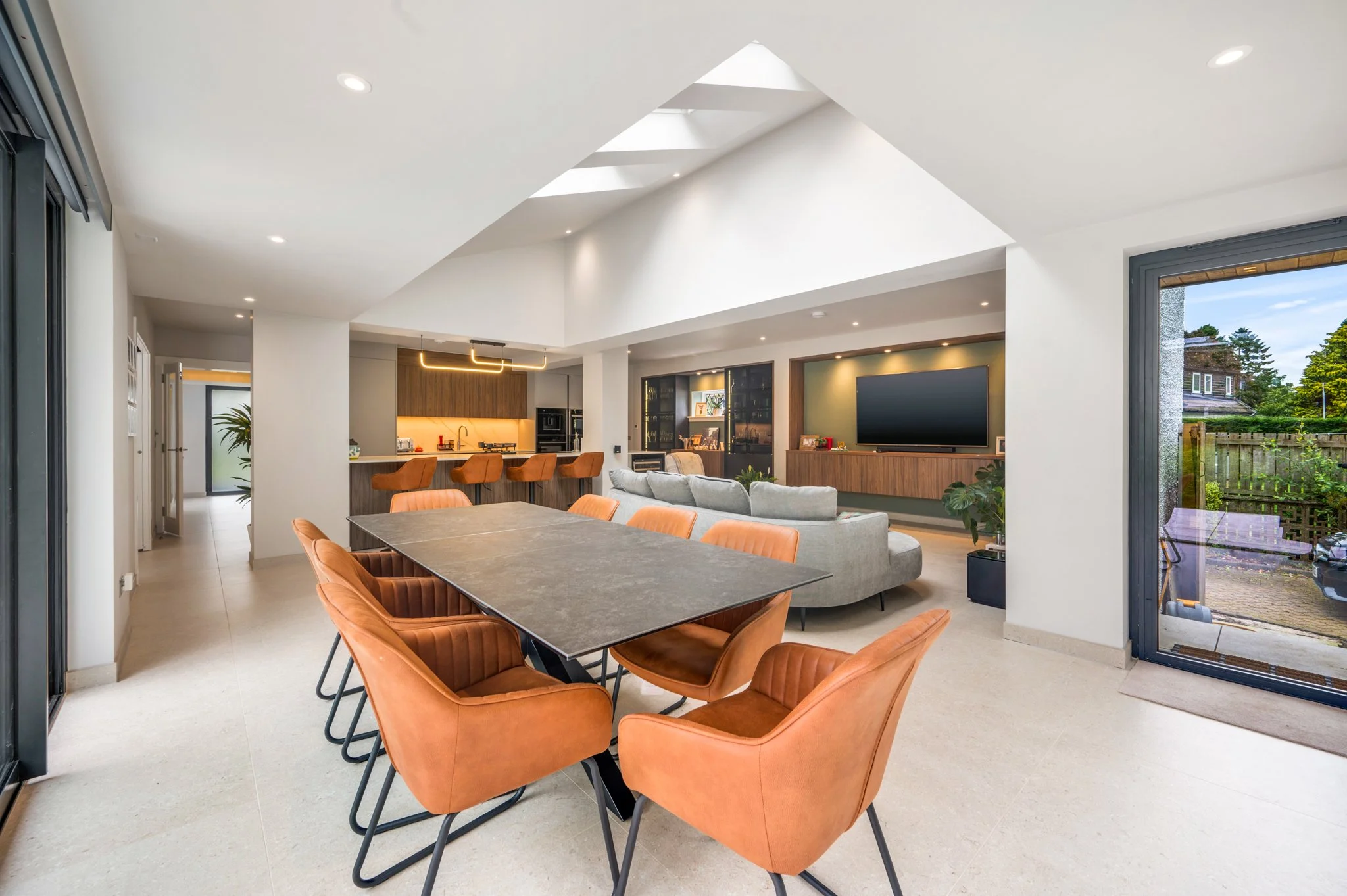 Modern open-plan home interior with dining table, orange chairs, kitchen, gray sectional sofa, and wall-mounted TV