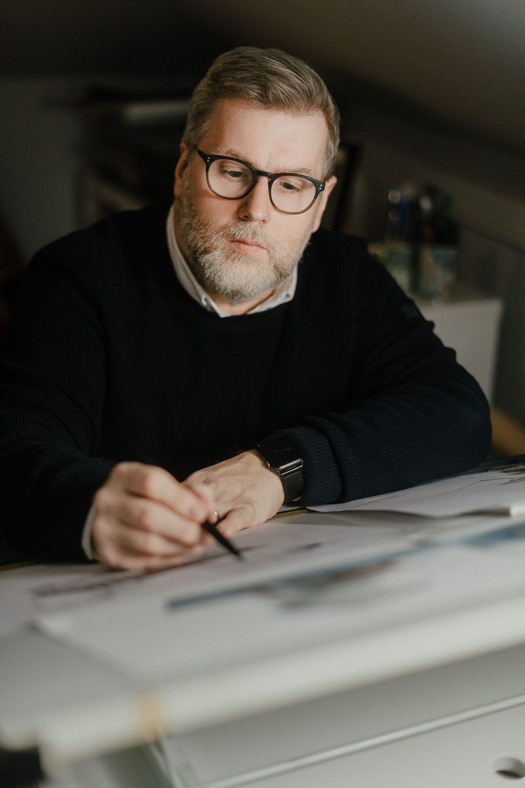 A man with glasses and a beard, wearing a black sweater and white shirt, sitting at a desk, writing or drawing on large sheets of paper.