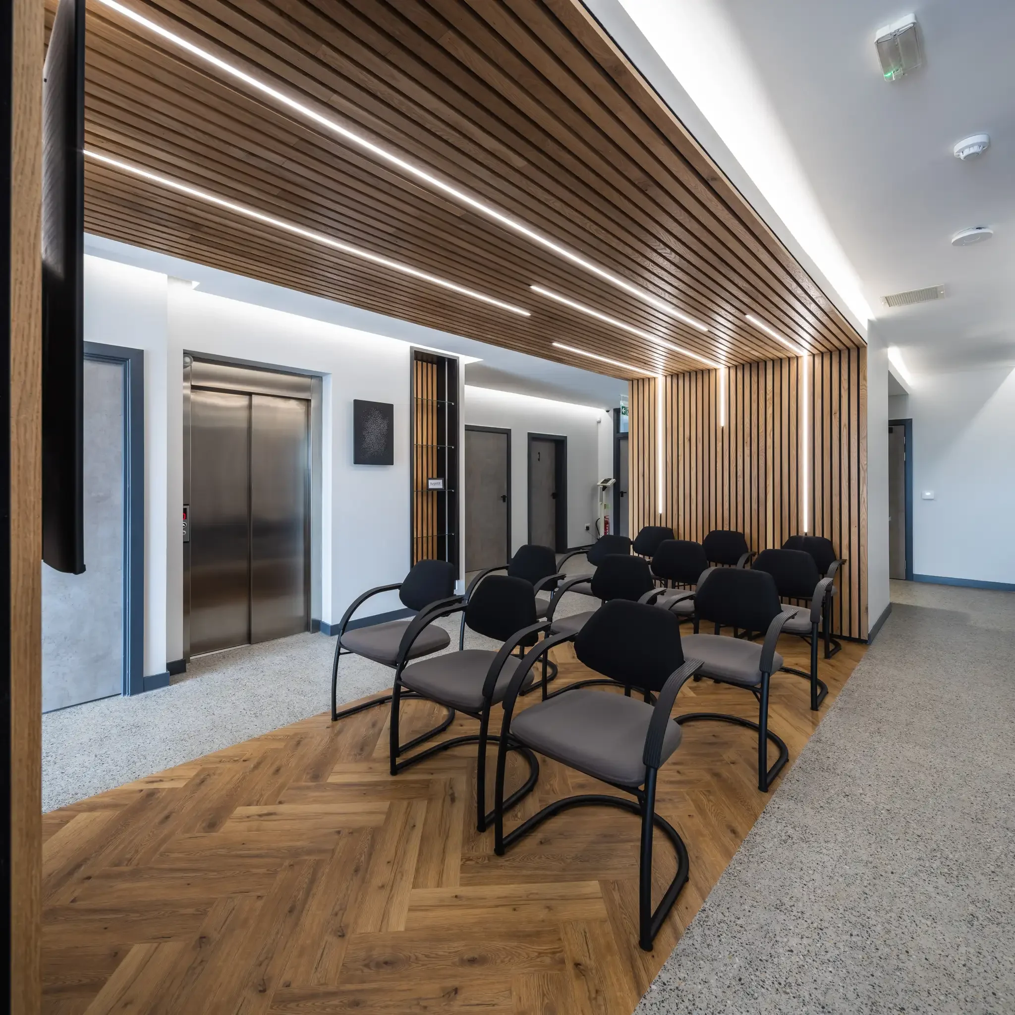 Modern waiting area with black chairs, elevator, wood-paneled ceiling and walls, and herringbone flooring