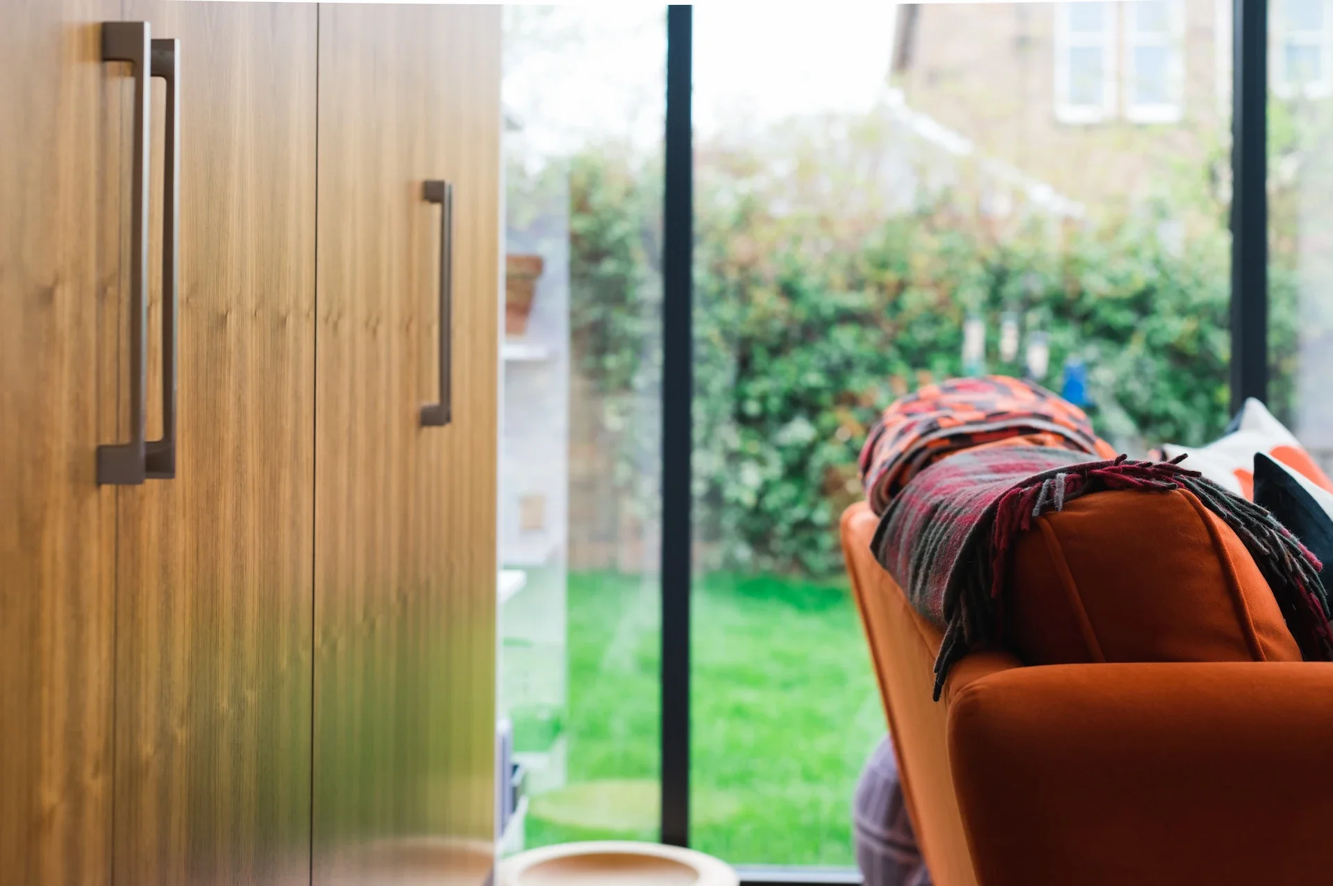 Wooden kitchen cabinets beside a person sitting in an orange armchair with a blanket, overlooking a garden through large windows