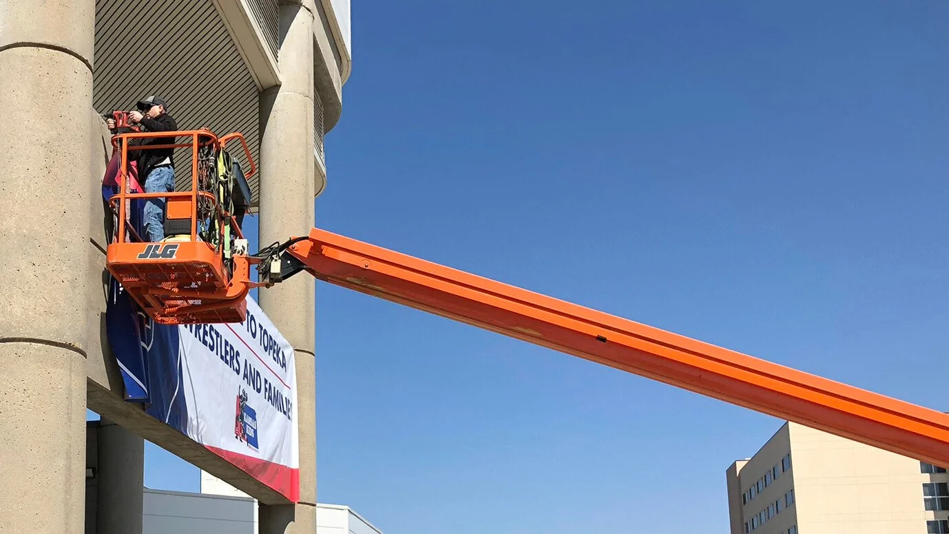 A worker elevated on an orange cherry picker is attaching a large banner to the side of a building. The banner reads "TO TOPEKA, WRESTLERS AND FAMILIES," with a logo at the bottom. The building is made of concrete with rounded columns, and a cityscape with a tall building is visible in the background.
