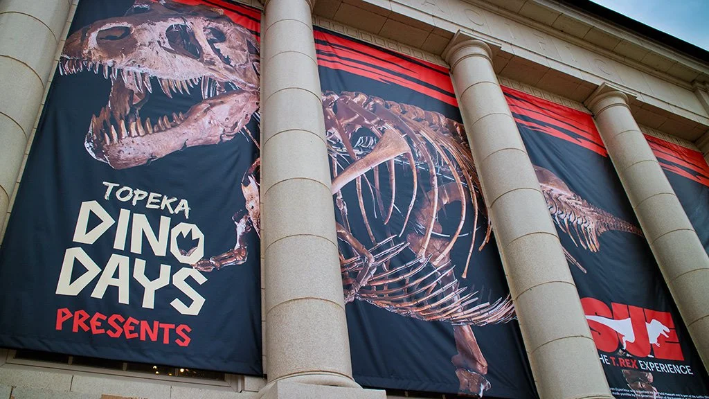 Large banners hanging on the front of a building, featuring a dinosaur skeleton and skull with the text "Topeka Dino Days Presents" and some red and white graphics representing T. Rex.