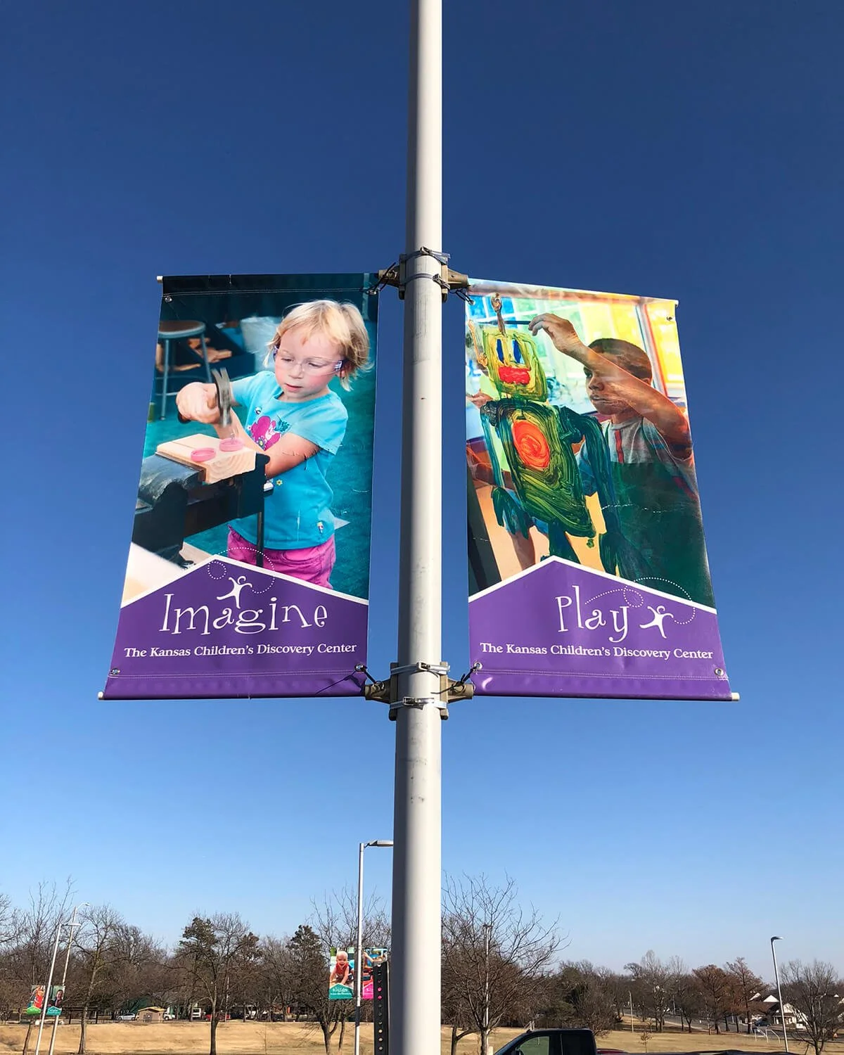 Two banners hanging on a light pole, one showing a young girl at a table with pink paint and the other showing a young boy painting a colorful picture, both at The Kansas Children's Discovery Center.