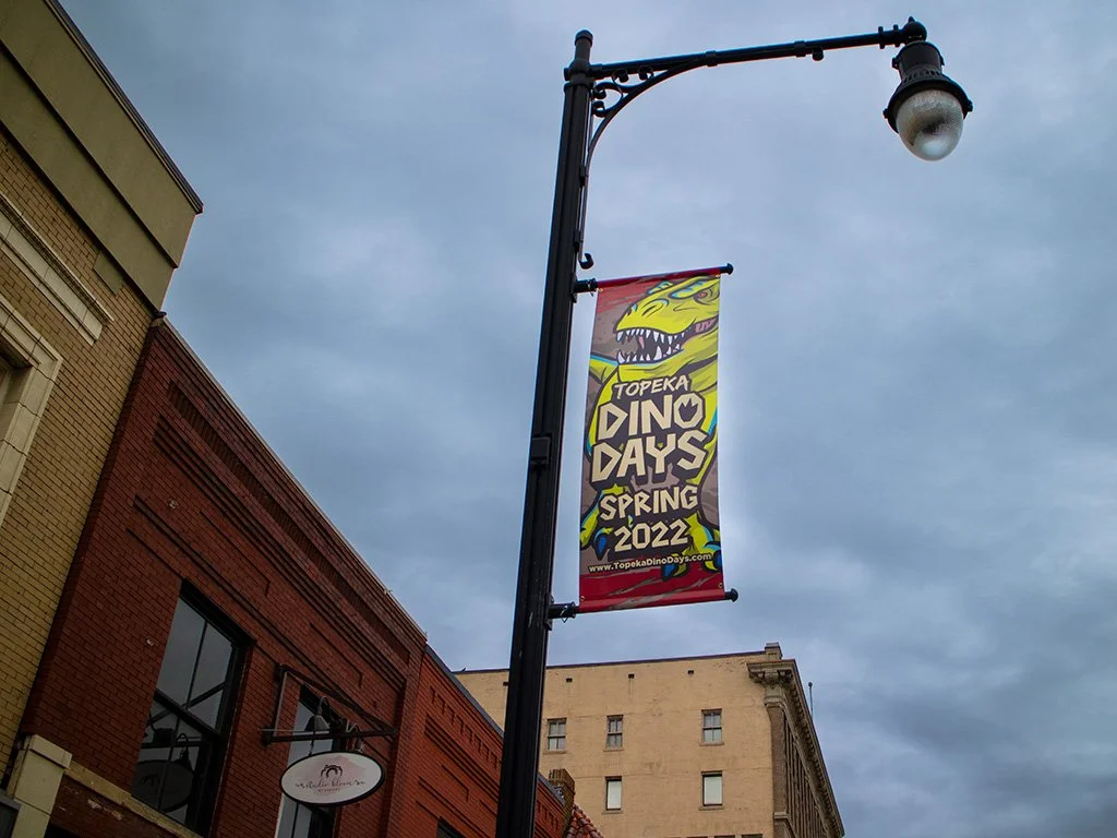A street lamp with a colorful banner advertising Topeka Dino Days Spring 2022, featuring a cartoon dinosaur head on a partly cloudy sky.