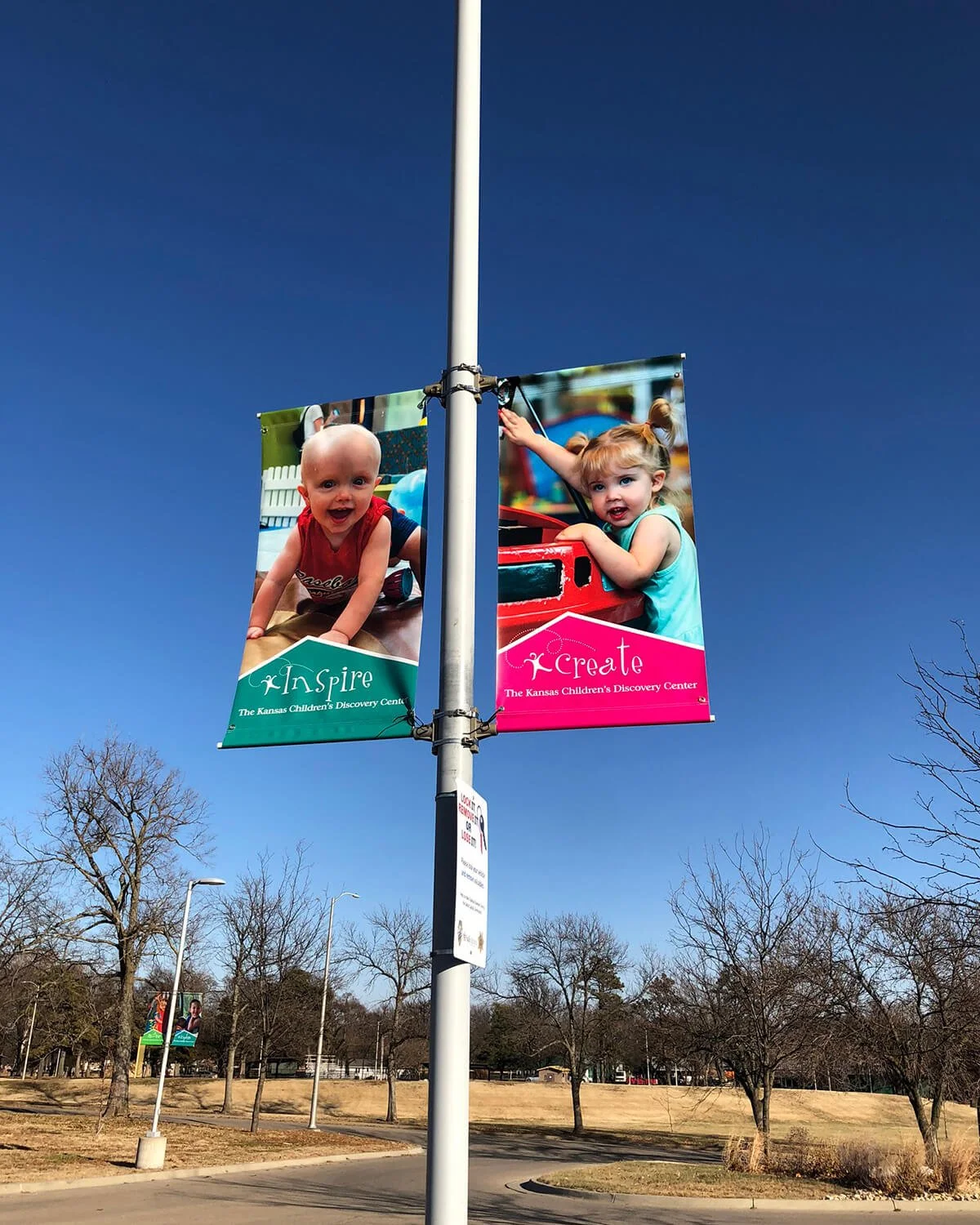 A street light with banners featuring two young children, a boy and a girl, at the Kansas Children's Discovery Center. The boy has a bald head and is smiling, while the girl with pigtails is leaning on a red toy. The banners are labeled "Inspire" and