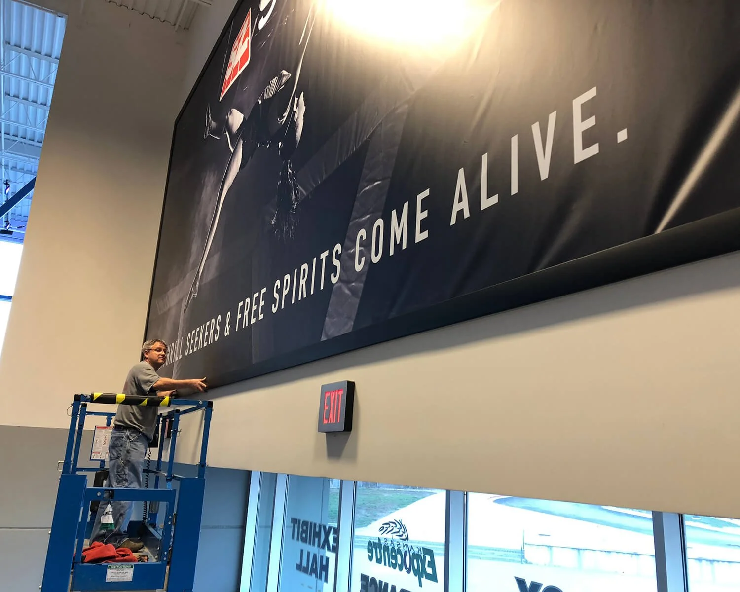 A man on a blue lift installing or adjusting a large billboard with the slogan 'Seekers & Free Spirits Come Alive' inside a building with high ceilings and large windows.