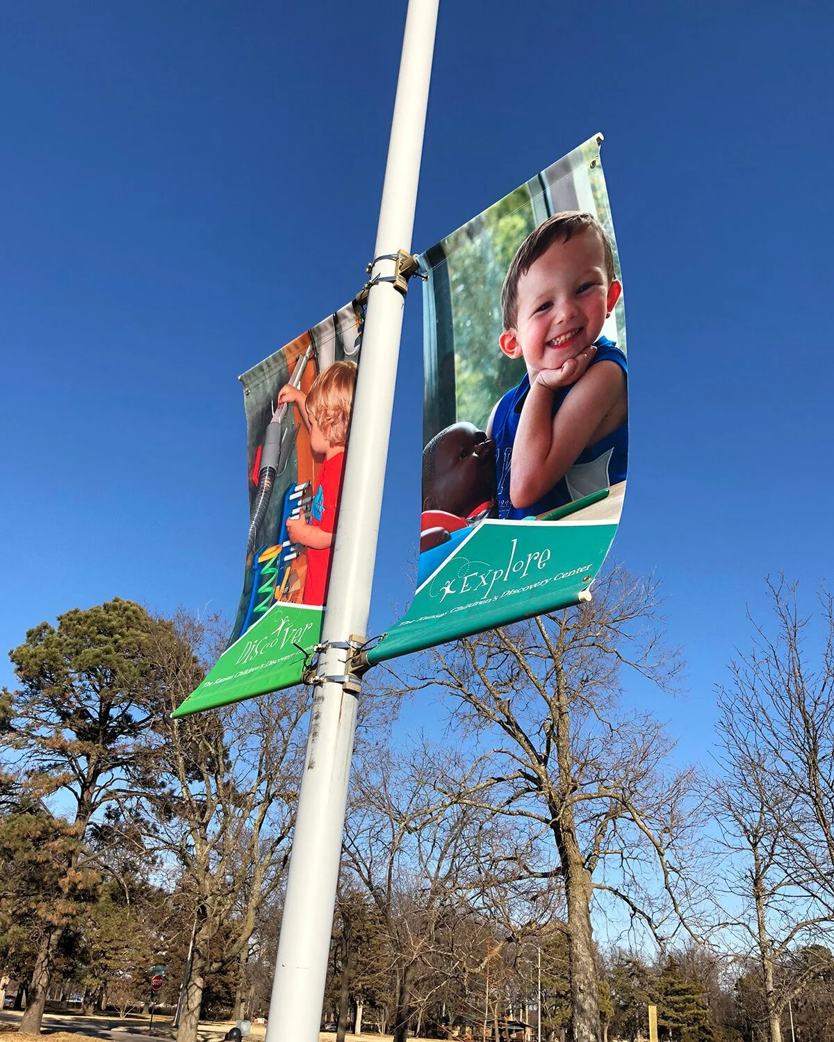 Banners hanging on a flagpole promoting the Kansas Children's Discovery Center, featuring images of children engaging in activities outdoors and indoors against a clear blue sky with trees in the background.