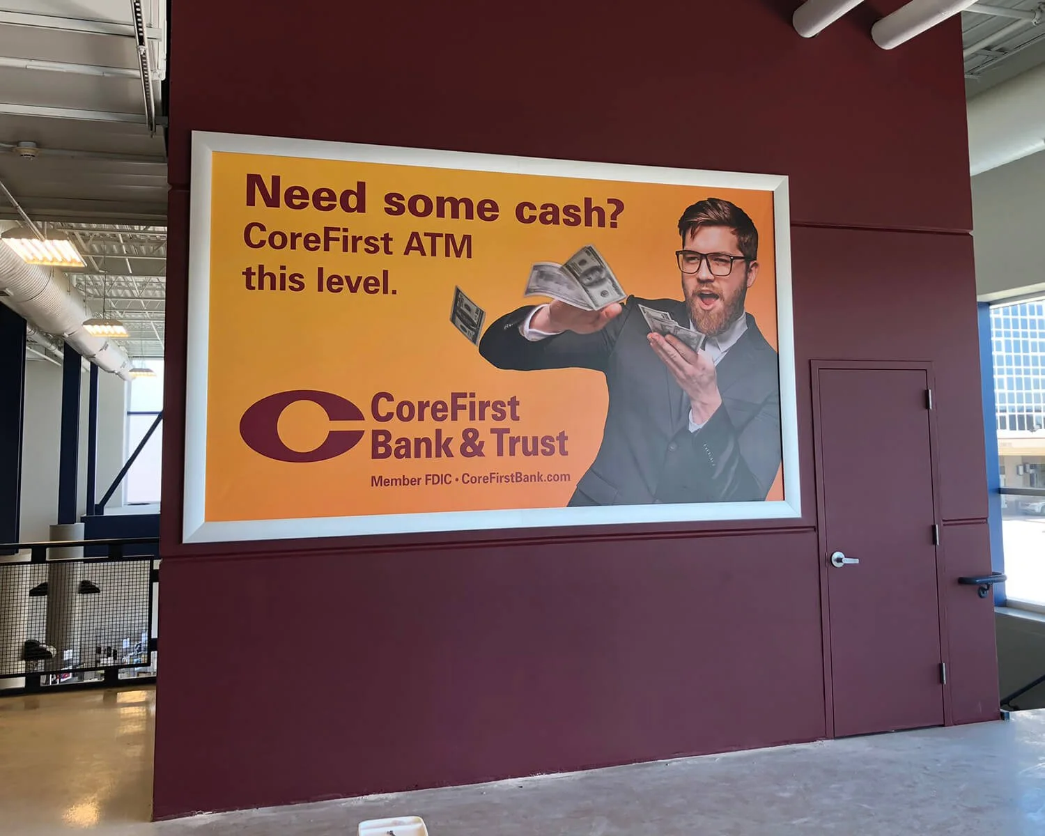A sign in an indoor space advertising CoreFirst Bank & Trust with a man in glasses holding cash with a surprised expression, and text that reads: 'Need some cash? CoreFirst ATM this level.'