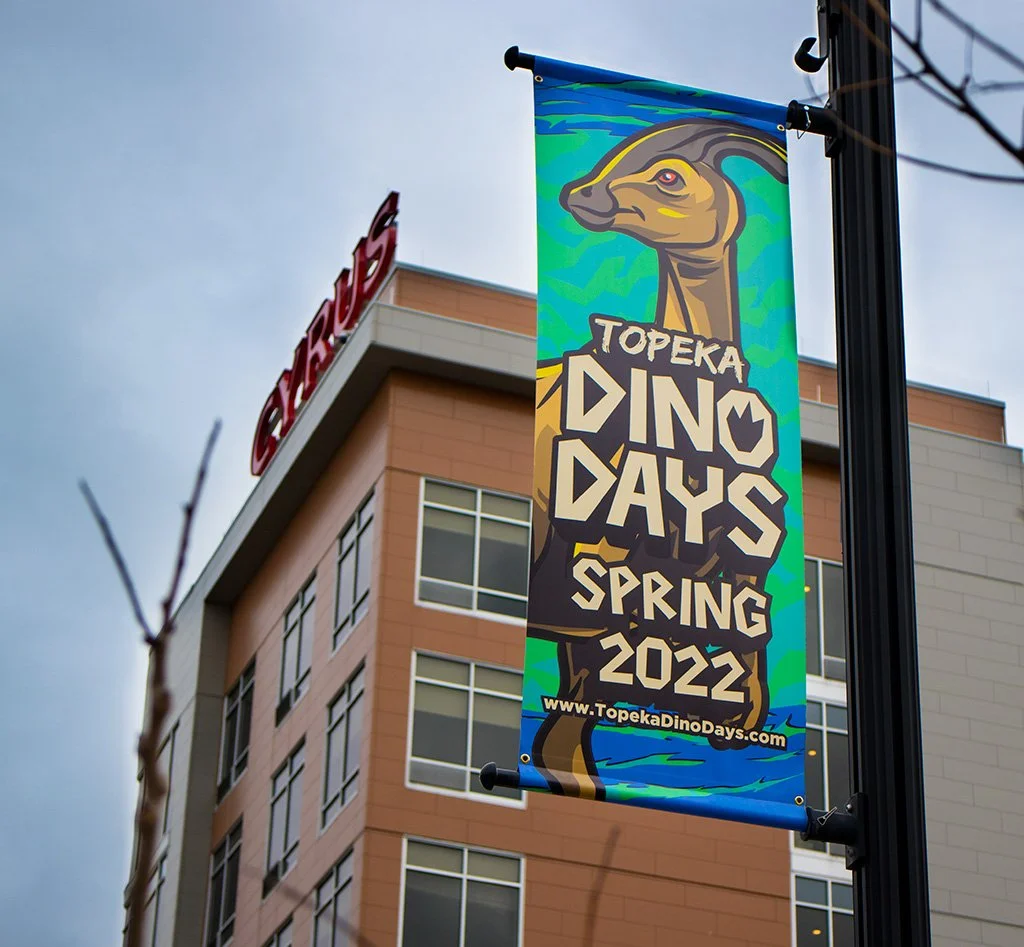 A colorful banner for Topeka Dino Days Spring 2022 hanging from a streetlamp with a building in the background.