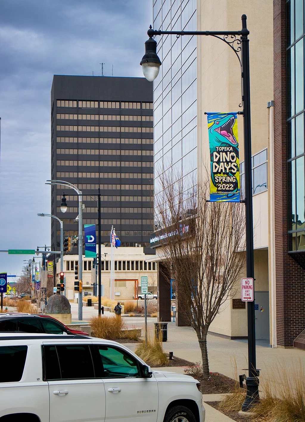 A city street with a banner reading 'Topeka Dino Days Spring 2022' hanging from a lamppost, buildings, parked cars, and pedestrians in the background on a cloudy day.