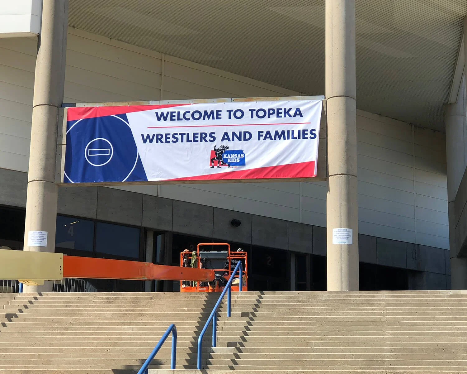 Banner hanging on the front of a building welcoming wrestlers and families to Topeka, Kansas. The banner has a sports theme with a hockey puck graphic and features the Kansas Kids logo.