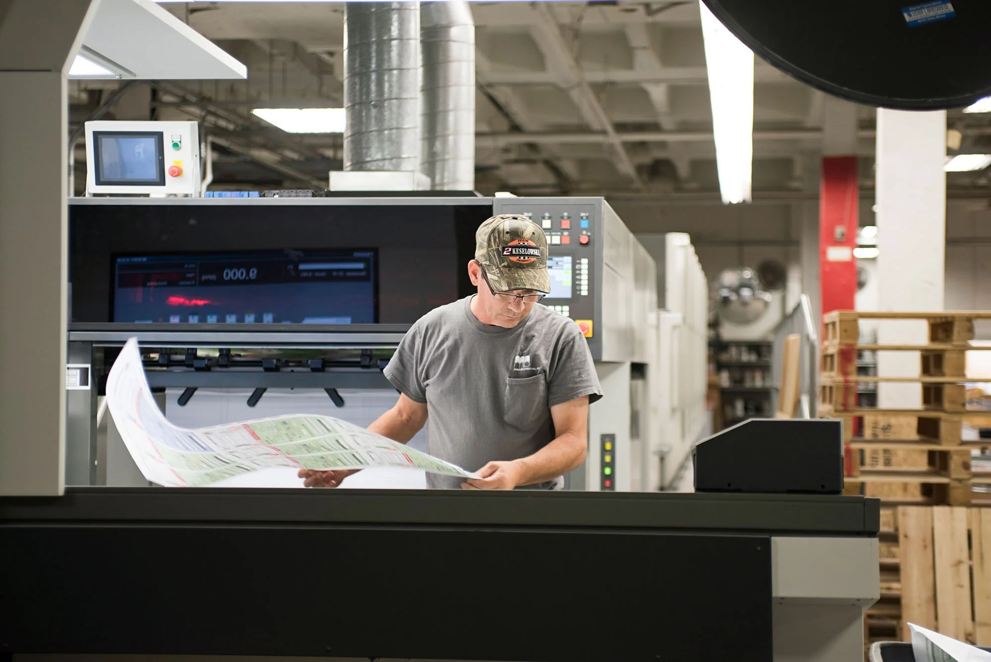 A man wearing a baseball cap and glasses inspects a large sheet of printed material in an industrial print shop, with printing equipment and wooden pallets in the background.
