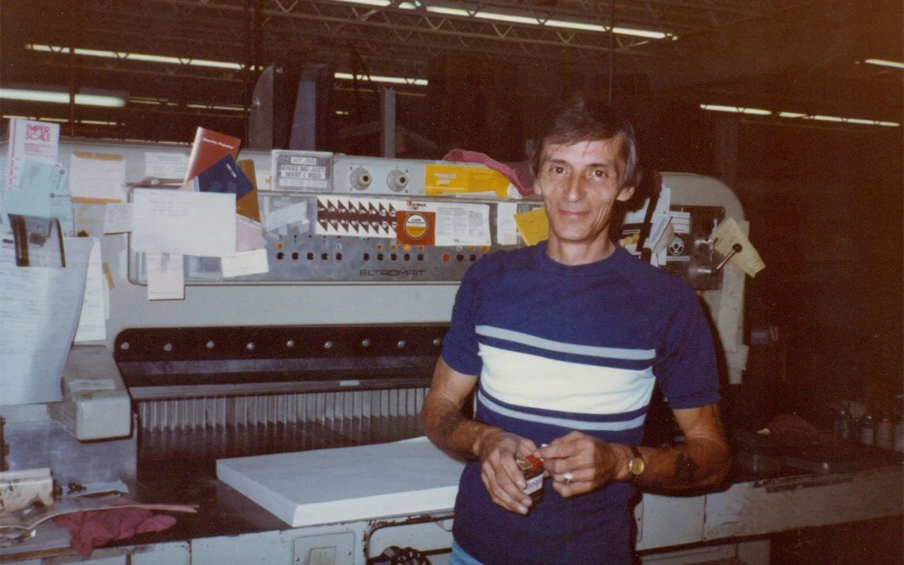 A man in a blue and white striped t-shirt standing in front of printing or packaging machinery in an industrial setting, holding a small object.