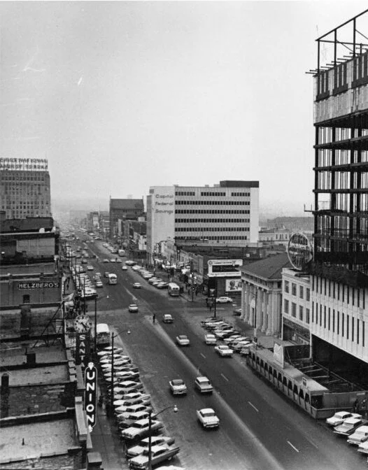 Black and white photo of a busy city street with cars parked along the sides and moving traffic. Buildings line both sides of the street, including high-rise and commercial structures. Some signage visible on the buildings includes a UNICO sign and a Helzberg sign.