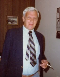 Man with gray hair wearing a dark blazer, tie, and light-colored shirt, holding a cigarette.