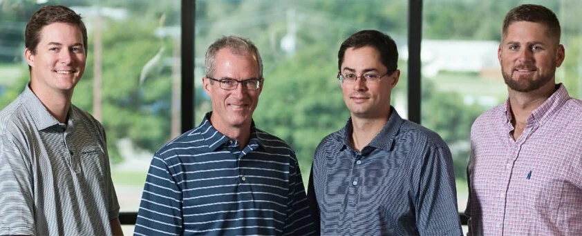 Four men standing side by side indoors with a large window and green scenery in the background.